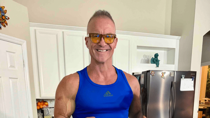 A medium shot of a muscular man in a kitchen wearing a blue Adidas tank top and yellow-tinted glasses. He is smiling at the camera; a refrigerator and kitchen cabinets are visible in the background.