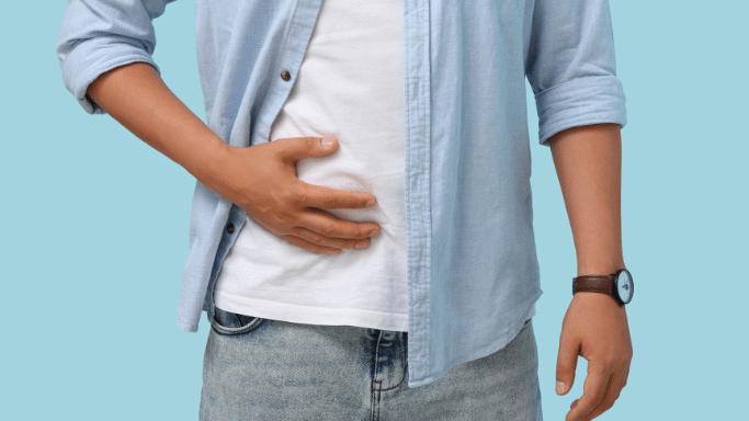 A close-up of a person's midsection against a light blue background. They are wearing a white t-shirt under a light blue button-down shirt and have their hand pressed against their stomach, suggesting abdominal pain or discomfort.