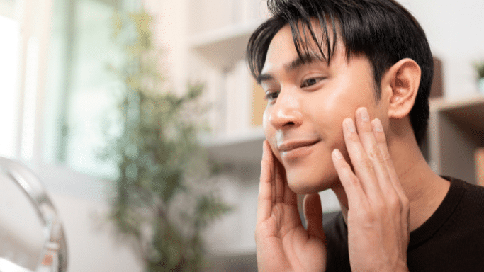 A close-up of a young man with dark hair looking into a mirror. He is gently touching his clean, clear skin on his cheeks with both hands, appearing satisfied with his complexion.