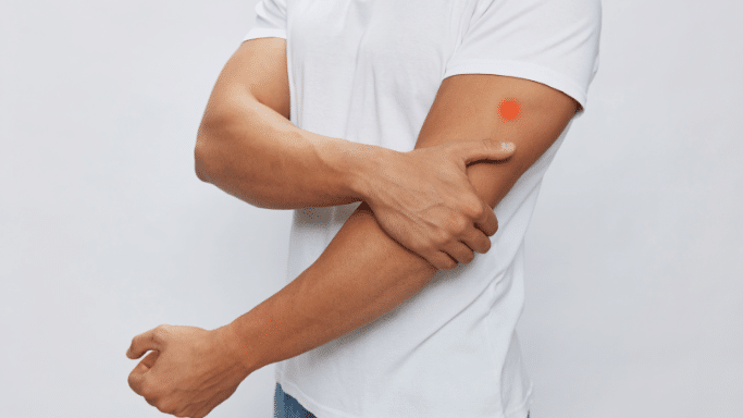 A man in a white t-shirt holding his upper arm. A glowing red circular spot is superimposed on his bicep to indicate a localized area of pain or an injection site.