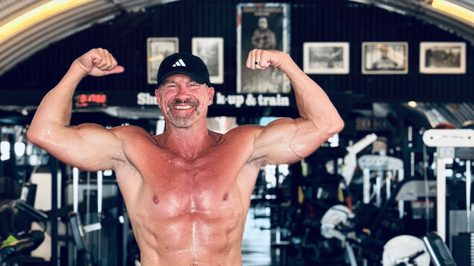 A middle-aged man with a goatee and a black Adidas baseball cap smiling broadly while flexing his biceps in a gym. He is shirtless, showing a muscular physique, with various gym equipment and framed photos visible in the background.