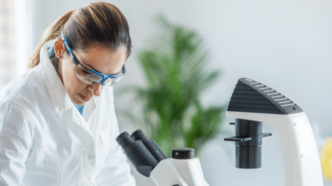 A female scientist wearing a white lab coat and blue-rimmed safety glasses looks down intently while working. Part of a modern microscope is visible in the foreground, with a blurred green plant in the background.