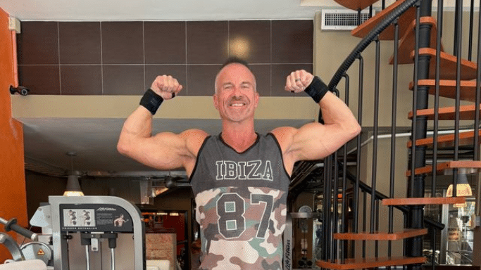A muscular man in a gray and camouflage "IBIZA 87" tank top smiles while flexing his biceps. He is standing in a fitness center near a Life Fitness cable machine and a wooden spiral staircase.