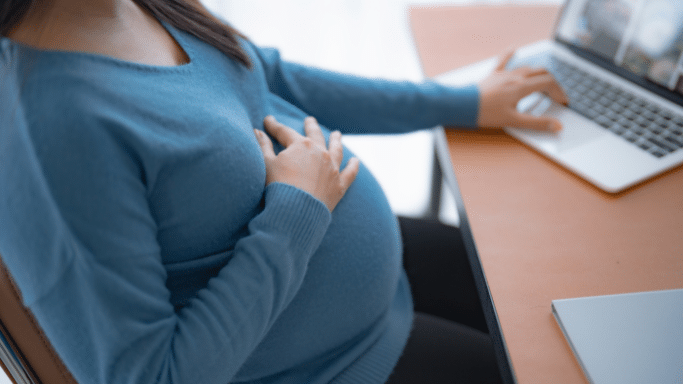 A side view of a pregnant woman sitting at a desk, wearing a blue sweater. One hand rests gently on her stomach while the other operates a laptop on a wooden surface.