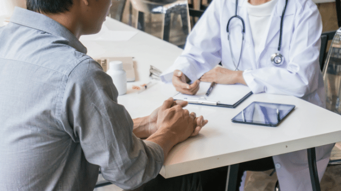 A person in a gray shirt sits across a desk from a doctor wearing a white lab coat and a stethoscope. On the desk between them are a tablet, a clipboard, a bottle of pills, and a syringe.
