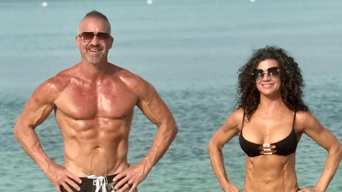 A man and a woman standing on a beach with the ocean in the background. Both are wearing swimwear and sunglasses, showcasing highly muscular, athletic physiques.