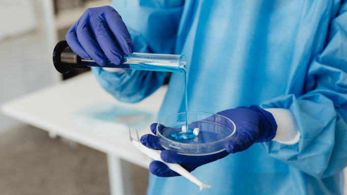 A close-up of a person in blue surgical scrubs and gloves pouring a bright blue liquid from a graduated cylinder into a petri dish containing small white tablets.