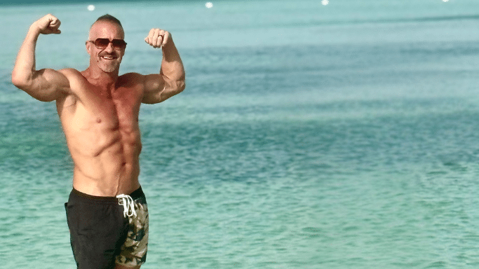 Man standing in the ocean and flexing his biceps. He is wearing camouflage-patterned swim trunks and sunglasses.