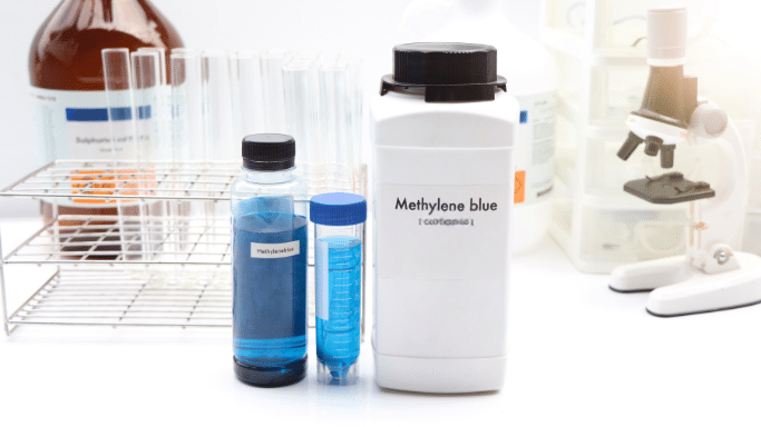 A close-up of lab equipment on a white surface. In the center is a large white container labeled "Methylene blue". Next to it are two smaller transparent bottles containing a vibrant blue liquid. The background features a rack of glass test tubes, a large amber chemical bottle, and a blurred white microscope.