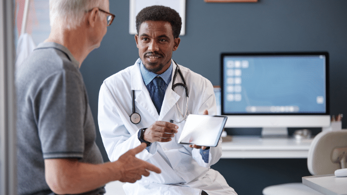 A medical consultation. A male doctor with a beard, wearing a white coat and a stethoscope, is sitting and holding a digital tablet. He is looking toward an older man (seen from the back) and appears to be explaining information displayed on the screen.