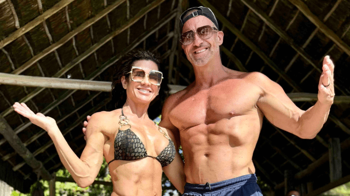 A man and a woman posing together outdoors under a thatched roof. Both are muscular and tanned, wearing sunglasses and swimwear. The man has his arm around the woman, and both are smiling broadly for the camera.