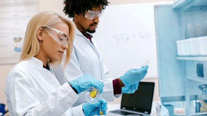 Two scientists working in a bright lab. A blonde woman in a white lab coat, safety glasses, and blue gloves is carefully pouring a yellow liquid into a test tube. Behind her, a male colleague is observing a sample.