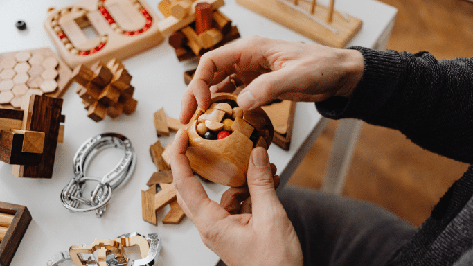 An overhead shot of a person's hands manipulating a complex wooden mechanical puzzle. The table is covered with various other wooden brain teasers, metal ring puzzles, and geometric shapes.