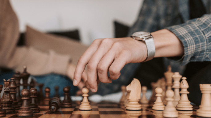A close-up, low-angle shot of a chess game in progress. A hand wearing a silver mesh-strap watch is reaching out to move a white pawn on the wooden board.
