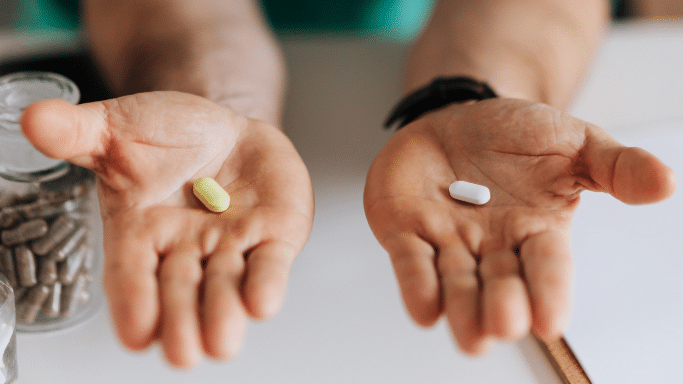 A first-person perspective of a person holding their hands open. In the left palm is a yellow pill, and in the right palm is a white pill. A bottle of capsules is visible on the table to the left.