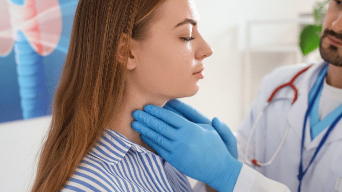 A close-up profile of a young woman undergoing a medical exam. A doctor wearing blue nitrile gloves is palpating her neck, likely checking the thyroid gland, while a medical illustration of a thyroid is visible in the background.