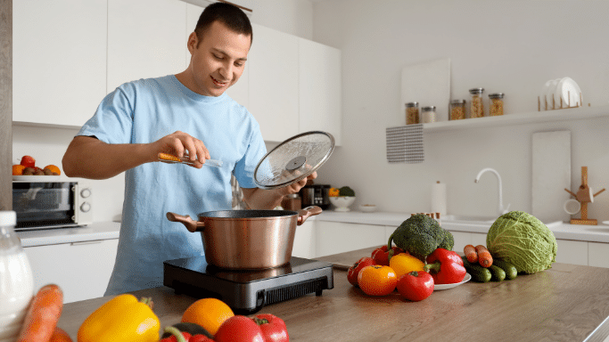 A smiling man in a light blue t-shirt stands in a bright, modern kitchen. He is holding a glass lid over a copper-colored pot on an induction cooktop, while adding spices from a small glass vial. The wooden counter in the foreground is filled with a vibrant variety of fresh vegetables and fruits, including peppers, tomatoes, broccoli, and carrots.