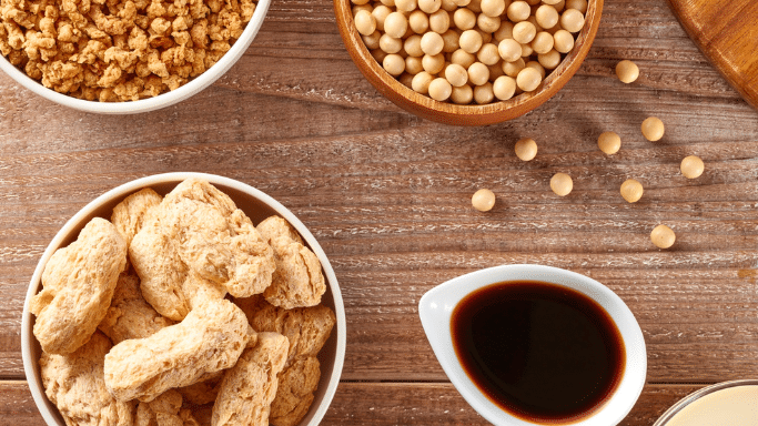 An overhead shot of various soy-based products on a wooden surface. There are white and wooden bowls containing dried soy chunks, whole soybeans, and soy granules. A small white pourer filled with dark soy sauce is also visible. An example of foods that lower testosterones levels