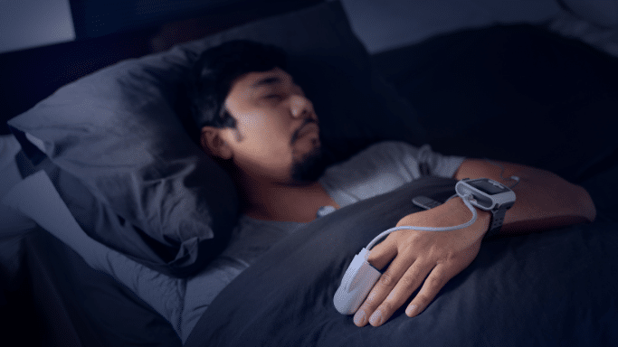 A man sleeping in a dark room wearing home sleep study sensors on his finger, wrist, and chest.