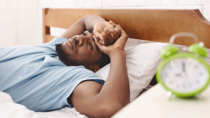 A man lying awake in bed with a frustrated expression; a blurred green alarm clock sits in the foreground.