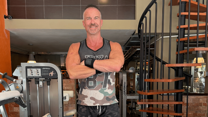 A muscular middle-aged man in a gym standing with his arms crossed and smiling.
