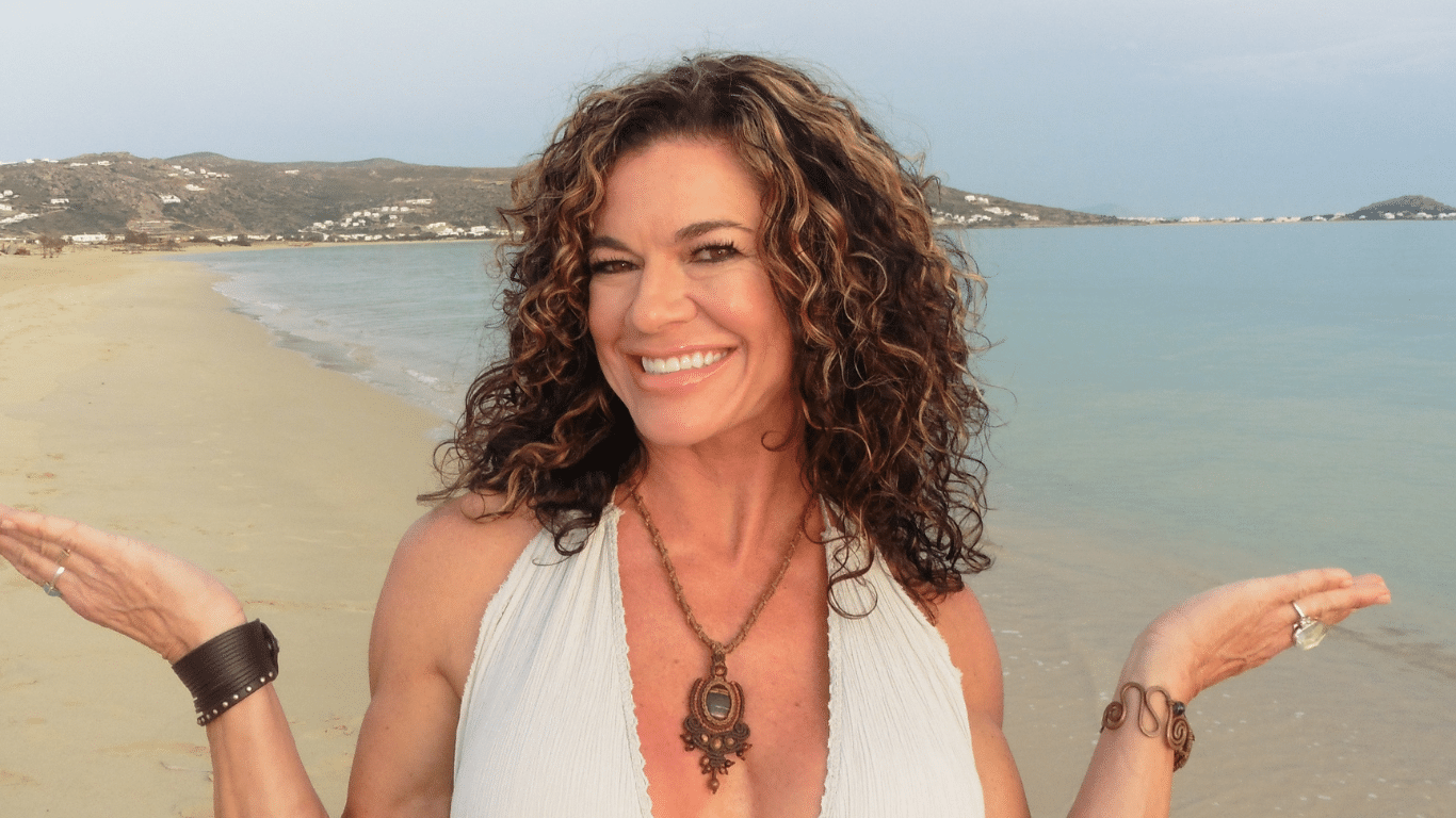 smiling woman with curly hair on a beach, wearing a white halter top and boho jewelry.