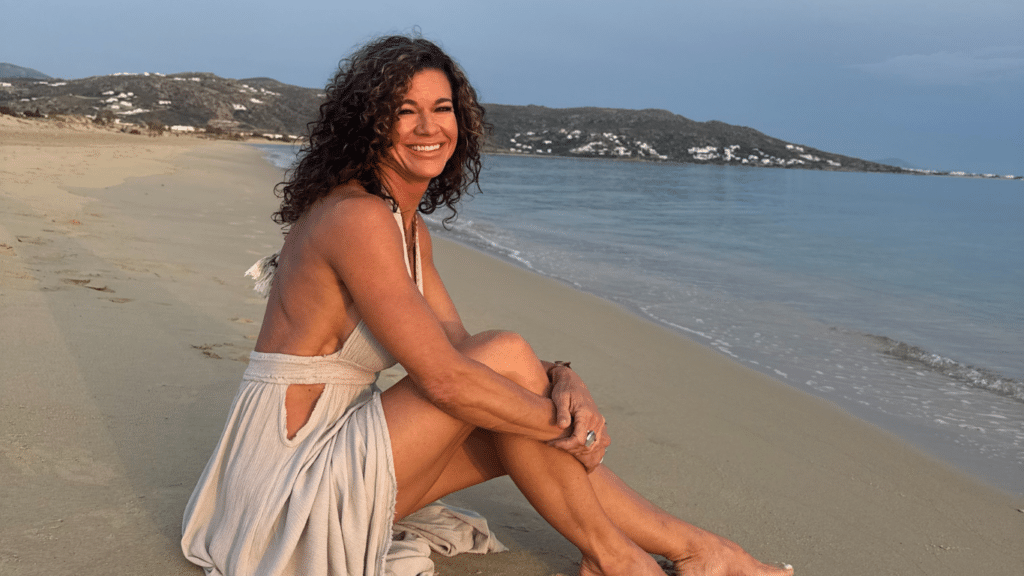 A woman in a white dress sitting on the sand at a beach, smiling toward the camera.