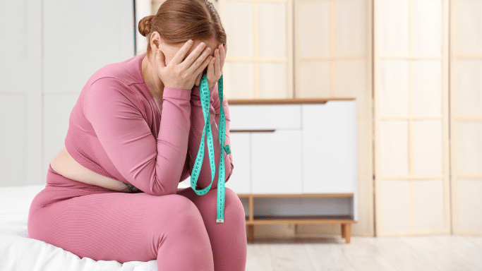 Person in pink activewear sitting on a bed, head in hands with a measuring tape