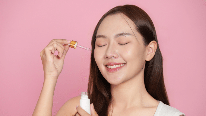 Woman smiling with eyes closed, applying facial serum with a dropper.