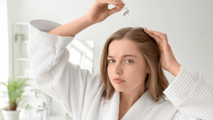 A woman in a white waffle-knit robe applying a treatment to her scalp with a dropper.