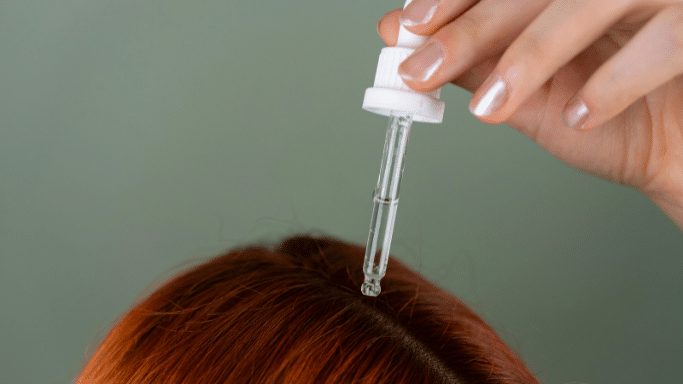 Close-up of a glass dropper releasing a clear liquid onto the part of a person's red hair.