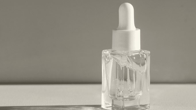 A square glass dropper bottle with clear liquid against a neutral, shadowed background.