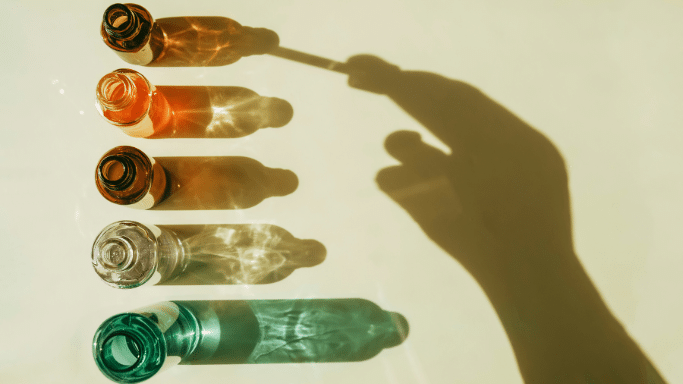 Top-down view of five colorful glass bottles casting long shadows, with a hand's shadow holding a dropper.