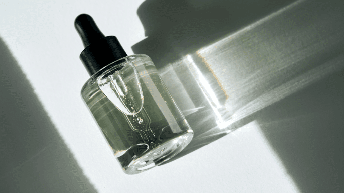 A round glass bottle with a black dropper cap laying on a white surface with sharp geometric shadows.