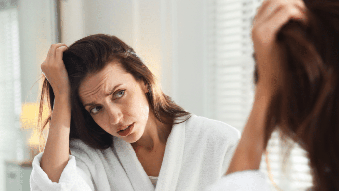 A woman with long brown hair looks into a bathroom mirror with a concerned and frustrated expression, running her hands through her hair to inspect her scalp.