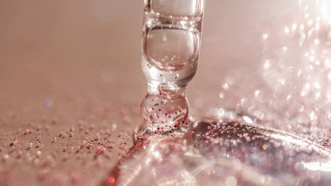 A macro shot of a glass pipette dispensing a thick, clear gel-like serum onto a surface. The serum contains fine, sparkling pink glitter.