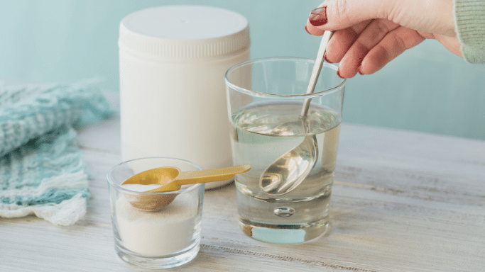 A person’s hand stirs a clear liquid in a glass with a spoon. On the table sits a large white supplement tub and a small glass containing white powder with a gold measuring scoop.
