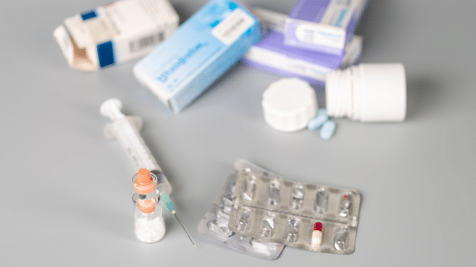 A high-angle, slightly blurred shot of various medical supplies scattered on a grey surface. Items include a syringe with an orange cap, several blister packs of pills (one containing a single red and white capsule), a small glass vial, and multiple blue and purple medication boxes in the background.