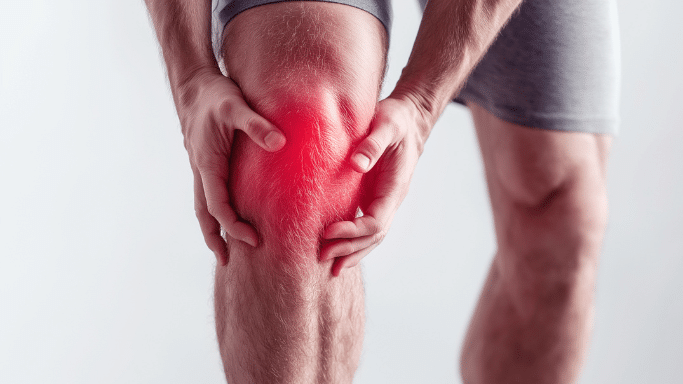 A close-up of a person in grey shorts holding their knee with both hands. A vibrant red, diffused glow is centered on the kneecap, suggesting intense localized pain or inflammation against a bright, clean background.