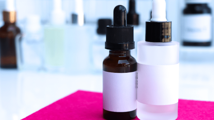 A close-up shot of two small dropper bottles standing on a bright pink surface. The bottle on the left is made of amber glass with a black dropper cap, while the bottle on the right is frosted glass with a white dropper cap. Both have blank white labels.
