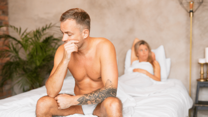 A man sits on the edge of a bed, looking down with a hand over his mouth in a pensive or distressed expression. In the background, a woman lies in the bed, looking toward him with a concerned or questioning look.