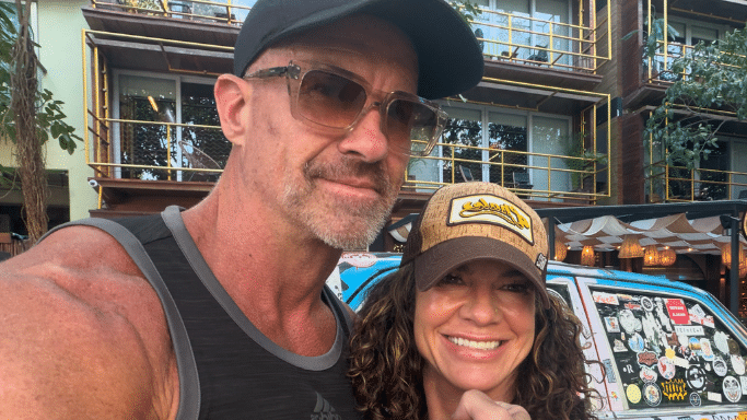 A close-up selfie of the same fit couple from image 382. The man is wearing sunglasses and a black cap, and the woman is wearing a brown trucker hat and smiling brightly.