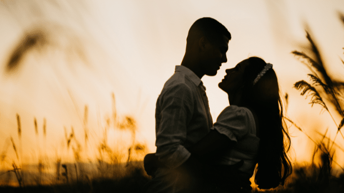 A silhouette of a couple embracing in a field during sunset. They are facing each other, leaning in close as if about to kiss, against a warm, golden-orange sky.