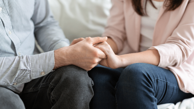 A close-up, shallow-focus shot of two people's hands clasped together. One person is wearing a striped shirt and the other a pink blazer, suggesting a moment of support or comfort.