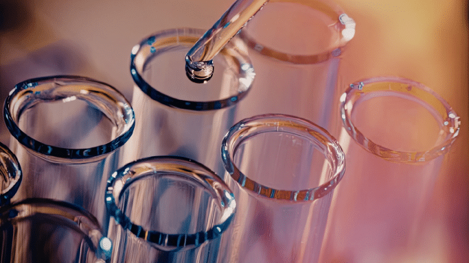 A close-up, artistic shot of several glass test tubes. A glass dropper is hovering over one of the tubes, with a single droplet of clear liquid about to fall. The lighting is warm and colorful.