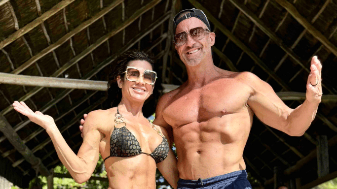 The fit couple from previous images poses outdoors under a thatched roof. The man is shirtless, showing a defined physique, and the woman is wearing a bikini. Both are wearing sunglasses and smiling with their arms outstretched.