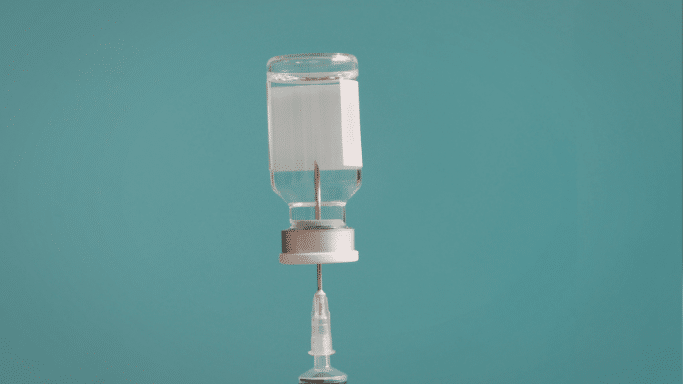 A medical glass vial turned upside down with a syringe needle inserted through the rubber stopper, ready to draw liquid against a plain teal background.