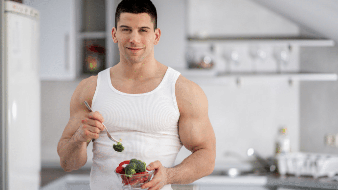 A muscular young man in a white tank top standing in a kitchen, holding a small glass bowl of fresh broccoli and red peppers, looking directly at the camera with a gentle smile.