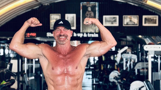 A close-up of the middle-aged man in a gym, shirtless and sweating, flexing his biceps with an intense, joyful expression.