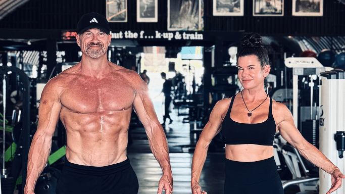 The muscular man stands next to a fit woman with her hair in a bun; both are in gym attire and posing in a weight room.